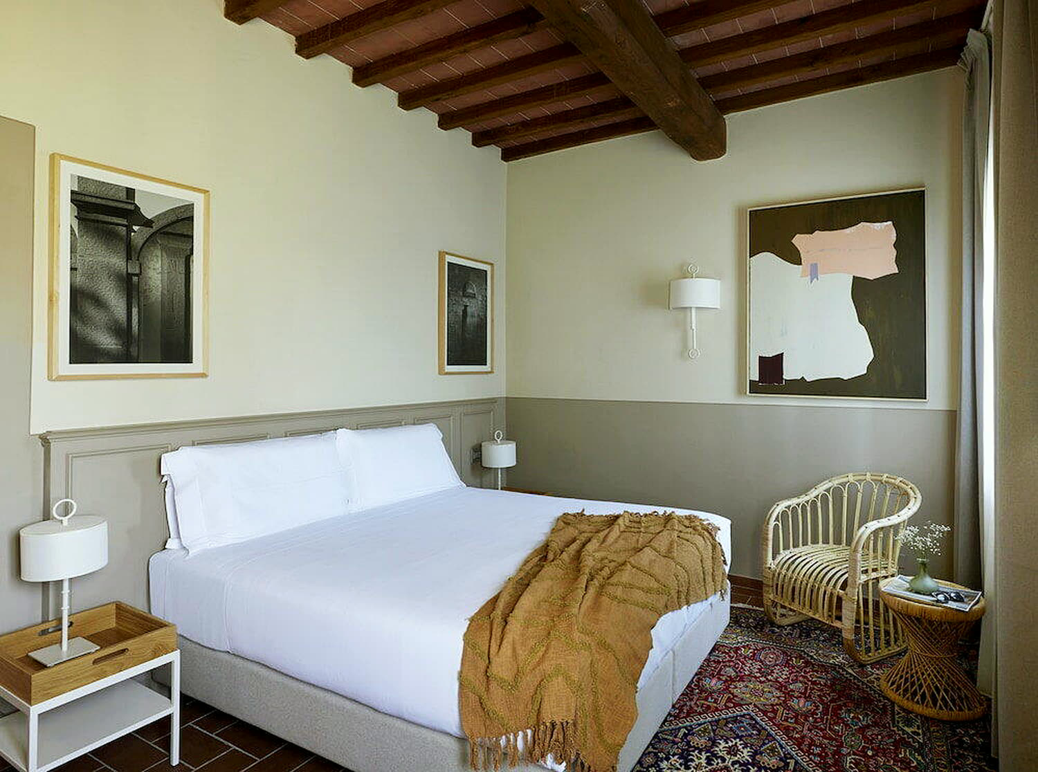 Camera con travi in castagno a vista, pareti chiare, letto in lino bianco e poltroncina in midollino.