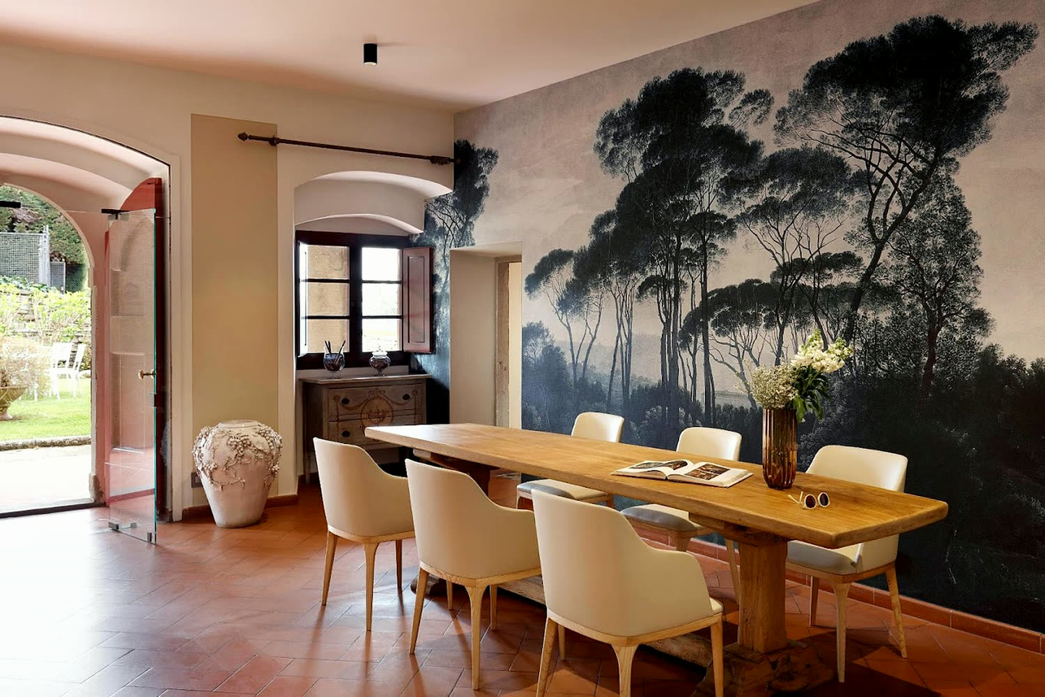 Sala da pranzo della tenuta: tavolo in legno massello, sedie chiare, pavimento in cotto e una parete dipinta con pini marittimi.